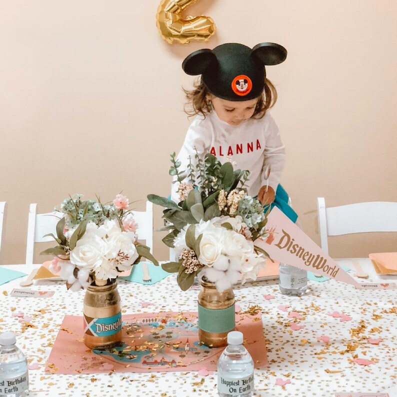 Puede incluir: Una mesa preparada para una fiesta de cumplea&ntilde;os con tem&aacute;tica de Disney World. La mesa est&aacute; cubierta con un mantel blanco con confeti dorado. Hay dos centros de mesa florales en tarros de vidrio con detalles dorados y turquesa. Los tarros est&aacute;n etiquetados con "Disney". Un bander&iacute;n rosa y azul con el texto "Disney World" est&aacute; sobre la mesa.