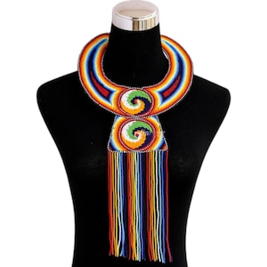 Puede incluir: Un collar de cuentas vibrante con un esquema de colores arcoíris. El collar presenta un gran collar curvo y un colgante central con diseños en espiral. Flecos de cuentas de colores cuelgan del colgante. Se muestra en un maniquí negro.