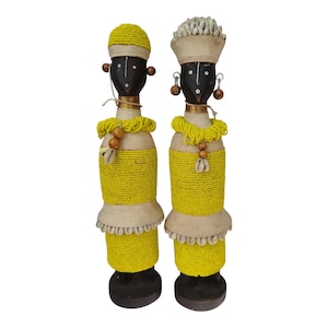 Puede incluir: Dos figuritas de madera de personas con caras negras, que llevan ropa y sombreros de cuentas amarillas y blancas. Las figuritas están de pie sobre bases de madera.