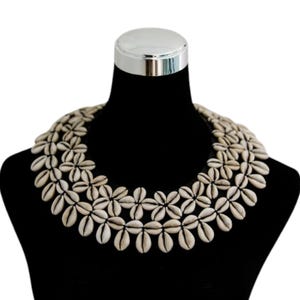 Op de afbeelding: Een opvallende ketting bestaande uit talrijke witte schelpen gerangschikt in een bloemmotief. De ketting wordt gepresenteerd op een zwarte mannequin, die het gedetailleerde ontwerp en de natuurlijke uitstraling benadrukt. De schelpen zijn crèmekleurig.
