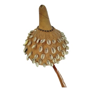 Puede incluir: Un instrumento de percusión artesanal, un agitador de calabaza, presenta un cuerpo bulboso adornado con numerosas conchas blancas. El cuello de la calabaza es alargado y un fino tallo marrón se extiende desde la base. El instrumento está diseñado para música rítmica.