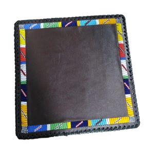 May include: Square, dark brown leather coaster with a beaded border. The border features a colourful pattern of blue, yellow, red, and white beads. The edges are stitched with black thread.