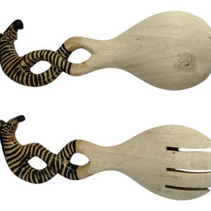 Op de afbeelding: Een paar houten saladeservers. De handvatten hebben een zebra-ontwerp met zwarte en bruine strepen. Eén server is een lepel, de andere heeft sleuven om te serveren. De houten servers zijn licht van kleur.