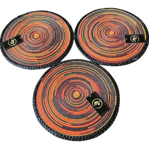 May include: Three circular placemats featuring a concentric pattern of orange, yellow, red, and black beads. Each placemat has a thick, braided black border and a small rectangular black tag with a gold circular logo attached to the surface.