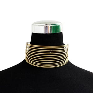 May include: A gold-colored choker necklace with multiple parallel wire strands. The necklace is displayed on a black mannequin, showcasing its design and form. The choker is a bold statement piece.