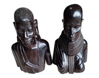 May include: Two dark brown wooden sculptures of human busts. The sculpture on the left has detailed facial features and a carved hairstyle. The sculpture on the right has a smooth head and a detailed collar. Both sculptures are on a white background.