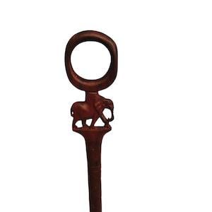 Puede incluir: Un abridor de botellas de madera marrón oscuro con un diseño de elefante tallado. El abridor tiene un mango circular y un cuerpo largo y delgado. El elefante se representa de perfil, de pie sobre una pequeña plataforma.