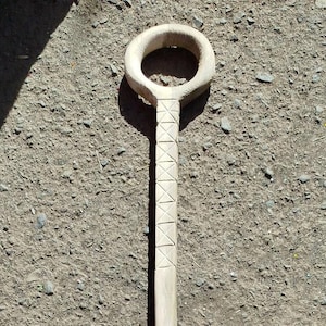 Puede incluir: Una herramienta de madera de color claro con un mango circular y un eje largo y rectangular. El eje presenta una serie de patrones tallados en forma de diamante. La herramienta probablemente se utiliza para jardinería o artesanía.