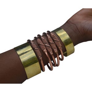Pode incluir: Um conjunto de cinco pulseiras, quatro são de fio de cobre com um design torcido e uma é uma pulseira larga de metal dourado.