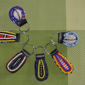 Pode incluir: Seis chaveiros de contas com suporte de couro e anéis de metal. Os chaveiros apresentam contas coloridas em vários padrões, incluindo um design de concha. Os chaveiros estão dispostos em uma superfície xadrez verde e bege.