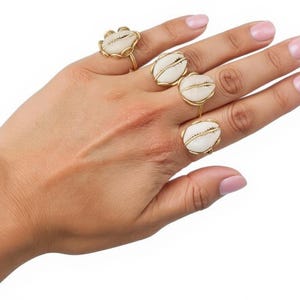 Puede incluir: Una mano que muestra cuatro anillos dorados con diseños de conchas blancas. Los anillos son de diferentes estilos, uno con forma de flor y los otros conchas ovaladas. Las joyas son de metal dorado.
