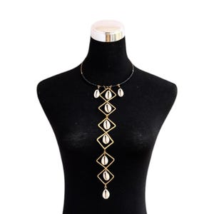 May include: A gold-coloured necklace with a geometric design, featuring diamond-shaped frames. Each frame holds a white seashell. The necklace is displayed on a black mannequin.