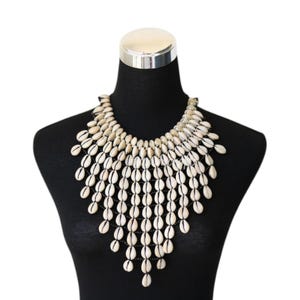 Op de afbeelding: Een opvallende ketting bestaande uit talrijke witte schelpen, gerangschikt in een trapsgewijs, gelaagd ontwerp. De schelpen zijn aan elkaar geregen, waardoor een textuurrijk accessoire ontstaat. De ketting wordt getoond op een zwarte mannequin.