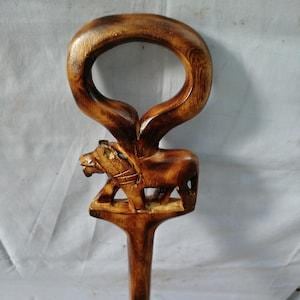 Puede incluir: Bastón de madera tallado a mano con una detallada escultura de un león. El león está posicionado sobre un pedestal, con un asa curvada en la parte superior. La madera tiene un acabado cálido y natural, mostrando la veta y la textura.