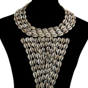 Op de afbeelding: Een opvallende ketting gemaakt van talrijke kleine, crèmekleurige kaurischelpen. De ketting is in meerdere strengen rond de hals gerangschikt en valt in een driehoekige vorm. De schelpen zijn aan elkaar geregen, wat een textuur en natuurlijke uitstraling creëert.