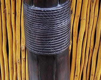 May include: A dark brown wooden sculpture of a human head and torso. The head has detailed facial features and a carved hairstyle. The neck is adorned with a carved band. The sculpture is set against a backdrop of vertical yellow bamboo.