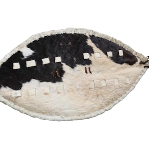 Puede incluir: Un escudo tradicional con un diseño único. El escudo presenta una combinación de piel de animal negra y blanca, con una serie de recortes rectangulares y elementos decorativos. Tiene una forma curva y un asa.