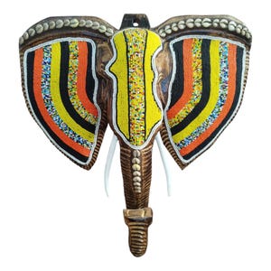 May include: A wooden elephant head wall hanging with colorful beaded accents. The elephant's ears are adorned with orange, yellow, black, and blue beads. The tusks are white, and the trunk is brown with a decorative pattern.