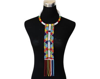 May include: A colorful beaded necklace with a white collar and a long, beaded pendant. The pendant features a geometric design in red, yellow, green, blue, and white beads, with a fringe of colorful strands.
