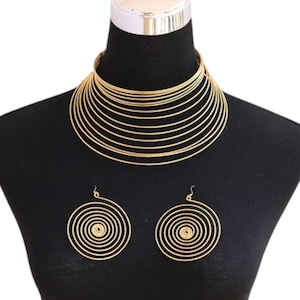 May include: Gold-coloured necklace and earrings set. The necklace has multiple stacked, circular bands, forming a choker. The earrings are spiral-shaped, mirroring the necklace. The jewellery is displayed on a black mannequin.