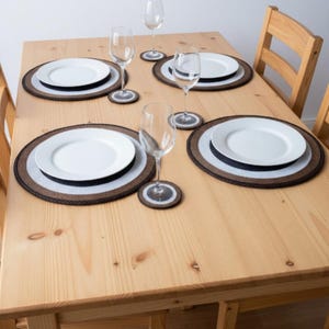 Könnte beinhalten: Ein Holztisch mit vier Gedecken. Jedes Gedeck umfasst einen weißen Teller, einen schwarzen Teller, eine braun-weiße Tischmatte, ein Weinglas und einen passenden Untersetzer. Der Tisch ist aus hellem Holz.