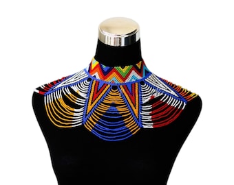 May include: A colorful beaded collar necklace displayed on a black mannequin. The necklace features a vibrant array of colors including red, yellow, blue, and white. The design incorporates geometric patterns and cascading beaded strands, creating a striking accessory.