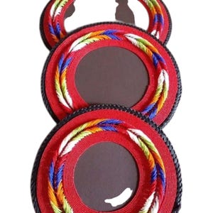 May include: Three circular red coasters with a dark brown center and a braided border featuring multicolored threads in blue, yellow, green, and white. The outer edge of each coaster is finished with a black braided trim, creating a textured appearance.