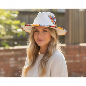 Op de afbeelding: Een witte fedora hoed met een kleurrijk bloemmotief aan de zijkant en een patroonrand. De rand van de hoed heeft een herhalend patroon van rood, oranje, geel, blauw en zwart. Een stijlvol accessoire.