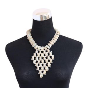 May include: A beige beaded necklace with a diamond-shaped pendant. The necklace is displayed on a black mannequin. The beads are oval-shaped and arranged in a layered pattern, creating a textured appearance. The pendant is a focal point.
