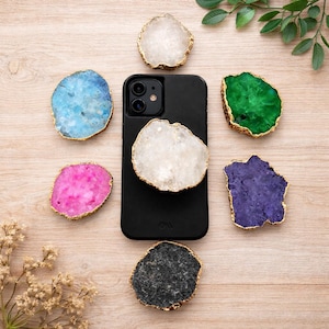 May include: A black phone case with a white crystal phone grip is surrounded by colorful crystal phone grips in blue, pink, green, purple, gray, and white. Each crystal has a gold-colored trim. The crystals are arranged on a light wood surface.