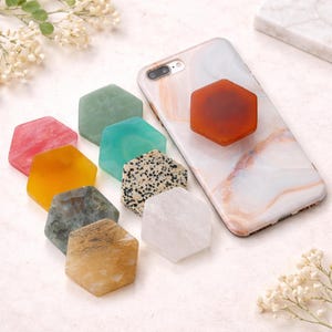 May include: A collection of hexagonal phone grips in various colors and textures, including pink, orange, green, and speckled stone. One grip is attached to a marble-patterned phone case. The grips are arranged on a white surface with floral accents.