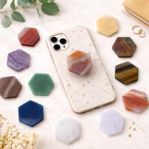 May include: A collection of hexagonal gemstone phone grips in various colors, including red, purple, green, and blue. One grip is attached to a white phone case. Other gemstones are scattered around the phone. The gemstones have unique patterns and textures.