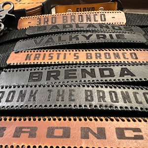 Puede incluir: Un conjunto de seis correas de cuero con diferentes nombres grabados en ellas, incluyendo "3rd Bronco", "Badlands Valkyrie", "Kristi's Bronco", "Brenda", "KRONK THE BRONCO" y "BRONCO".