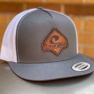 Peut inclure: Casquette de camionneur grise et blanche avec un patch en cuir marron qui dit "Outer Banks" et un motif de vague.