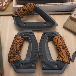 Puede incluir: Tres agarraderas automotrices de color gris oscuro con cubiertas de patrón topográfico naranja y negro. Cada agarradera tiene la palabra BRONCO impresa en negro sobre la cubierta. Las piezas presentan un diseño angular texturizado con orificios de montaje visibles.