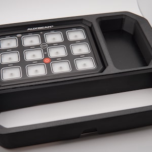 Könnte beinhalten: Schwarzes Bedienfeld mit dem Text "AUXBEAM" und mehreren quadratischen Tasten. Das Bedienfeld hat einen Ein-/Ausschalter und eine Reihe kleinerer Tasten. Das Bedienfeld ist in einem schwarzen rechteckigen Rahmen mit drei Fächern angeordnet.