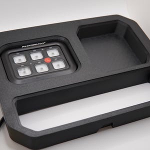 Könnte beinhalten: Schwarzes Bedienfeld mit dem Text "AUXBEAM" und sechs quadratischen Lichtern, einem roten Knopf und einer Ablage. Das Panel ist in einem schwarzen rechteckigen Rahmen mit einer zweiten Ablage angeordnet. Ein gelb-schwarzes Kabel ragt aus dem Rahmen.