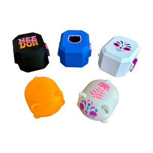Könnte beinhalten: Eine Sammlung von fünf kleinen Spielzeugen, bestehend aus drei achteckigen Behältern und zwei abgerundeten Figuren. Die Behälter sind schwarz mit dem Text NEE DOH, blau mit einer Katzenkontur und weiß mit einem bunten Katzengesicht. Zwei Figuren sind orange und weiß.
