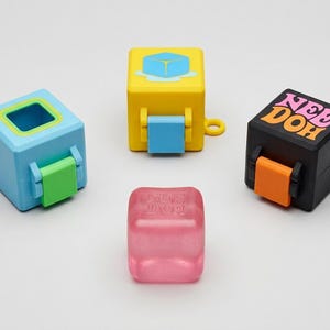 May include: Three square plastic containers and one translucent pink cube. The light blue container has a green latch, the yellow container features an ice cube graphic and blue latch, and the black container displays the text NEE DOH with an orange latch.