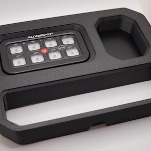 Könnte beinhalten: Schwarzes Bedienfeld mit dem Text "AUXBEAM" und beleuchteten Tasten. Das Panel ist in eine strukturierte schwarze Ablage mit zusätzlichen Fächern eingesetzt. Ein Netzschalter ist sichtbar. Ein schwarzes Kabel erstreckt sich von der Einheit.