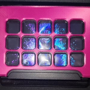 以下が含まれることがあります： ピンクのキーボードケースに、16個の黒いキーのグリッドがあります。各キーには、透明な青と黒のグラフィックデザインが施されています。キーの1つに「ITZ」というテキストが表示されています。