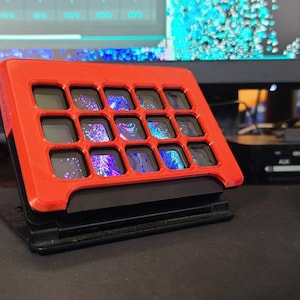 以下が含まれることがあります： 16個の小さな画面のグリッドを持つ赤と黒のキーボード。各画面には、異なるカラフルなパターンが表示されます。キーボードは黒い表面にあります。