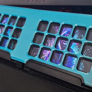 以下が含まれることがあります： 20個の正方形の開口部を持つターコイズブルーのキーボードケース。各開口部は、カラフルな抽象的なデザインを明らかにする透明なプラスチック製の窓を持っています。
