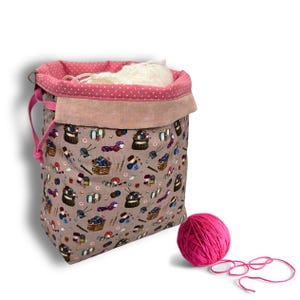 Könnte beinhalten: Ein Stoffbeutel mit Kordelzug in Beige mit einem wiederkehrenden Muster aus Wollkörben und Stricknadeln. Das Innenfutter ist rosa mit weißen Punkten. Ein einzelnes Knäuel aus leuchtend rosa Wolle liegt neben dem Beutel.