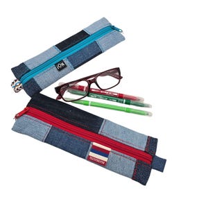 Könnte beinhalten: Zwei rechteckige Federmäppchen aus Jeansstoff im Patchwork-Design in Hell- und Dunkelblau. Eines hat einen hellblauen Reißverschluss und ein iON-Etikett, das andere einen roten Reißverschluss und ein Etikett mit der Aufschrift TESTSTREIFEN. Dazwischen liegen eine weinrote Brille und zwei Stifte.