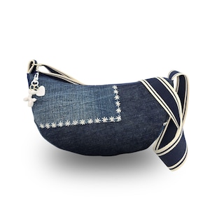 Könnte beinhalten: Eine Jeans-Umhängetasche mit geschwungener Form. Die Tasche hat weiße, gestickte Blumenakzente und einen gestreiften marineblauen und weißen Riemen. Ein kleiner Anhänger mit einem Herz und einem Bären hängt am Reißverschluss.