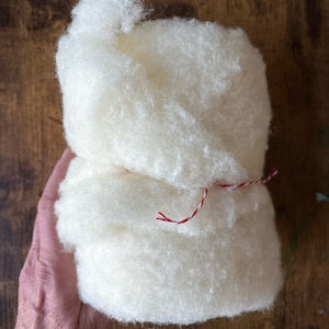 May include: A bundle of off-white, fluffy raw wool fiber with a soft, textured appearance. The cylindrical mass of wool is secured in the center with a thin, decorative string featuring a twisted red and white pattern tied in a simple knot.