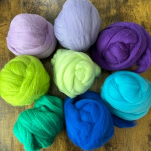 May include: Eight rounded bundles of soft, fluffy wool roving in a vibrant color palette. The collection includes shades of light purple, lavender, deep purple, lime green, bright yellow-green, emerald green, royal blue, and bright turquoise.