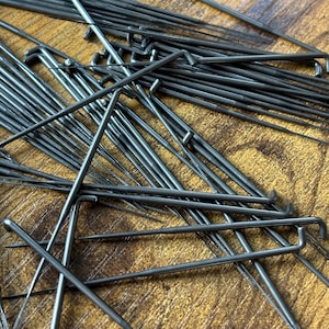 May include: A collection of numerous dark grey metal sewing machine needles, each featuring a straight shaft and a distinct right-angled bend at the top end. The needles are scattered in various directions, showcasing their sharp points and metallic finish.