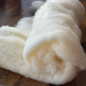 May include: A soft, off-white roll of raw wool fiber with a fluffy, fibrous texture. The material is loosely coiled, showcasing the natural, unspun state of the white wool. The fibers appear dense and airy, forming a continuous, thick layer.